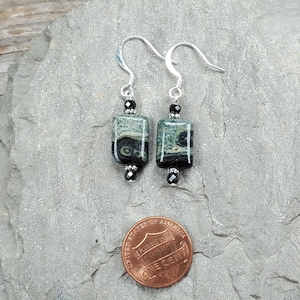 May include: A pair of dangle earrings with silver-tone hooks. Each earring features a rectangular, green and black stone bead, accented with black beads. The earrings are displayed on a gray, textured surface, with a penny for size reference.