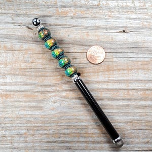 Puede incluir: Un bolígrafo estilete negro con una punta plateada y una sección de cuentas decorativas. Las cuentas son verdes, azules y doradas con un patrón floral. El bolígrafo mide aproximadamente 12,7 cm de largo y está sobre una superficie de madera.