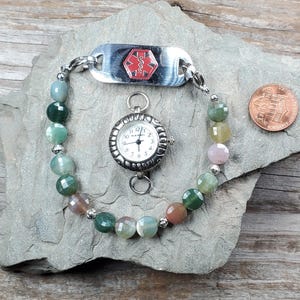 May include: A beaded bracelet with a silver medical ID tag featuring a red medical symbol. A small silver watch with a white face is attached. The bracelet has multicolored beads and a penny is next to it.