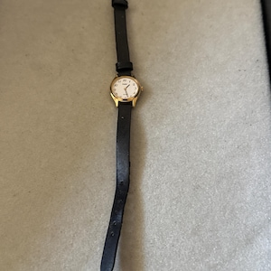 May include: A gold-coloured wristwatch with a white face and black numerals. The watch has a black leather strap with a silver buckle. A classic design, suitable for everyday wear.