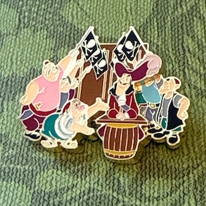 May include: A metal enamel pin featuring a group of cartoon pirates. One pirate stands behind a brown barrel, wearing a red coat and a purple hat. Other pirates in pink, blue, and white clothing surround the central figure, with two black flags featuring white skull and crossbones symbols displayed in the background.