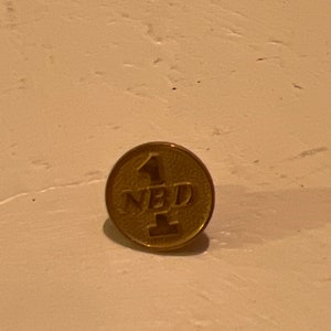 Może przedstawiać: Złota szpilka z wytłoczonym tekstem "1 NBD 1".