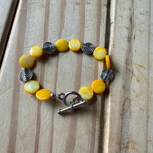May include: A bracelet featuring round, yellow, iridescent shell beads alternating with silver-toned metal beads embossed with a spiral pattern. The piece is secured with a silver-toned metal toggle clasp consisting of a circular ring and a T-bar.