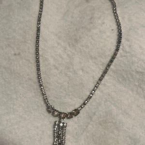 May include: A silver necklace with a dangling crystal pendant. The necklace is made of small, clear crystals that are set in a silver chain. The pendant is made of larger, clear crystals that are set in a silver frame.