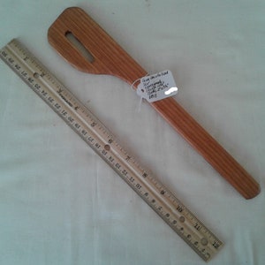 Könnte beinhalten: Ein Holzspatel mit einem Griff, der etwa 30 cm lang ist. Der Spatel hat ein kleines Loch oben und ist aus hellem braunem Holz gefertigt.