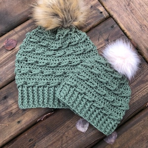 May include: A green crocheted hat with a brown and white pom pom and a matching green crocheted scarf with a white pom pom.