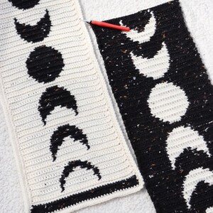 Puede incluir: Dos paneles de crochet con un dise&ntilde;o de fases lunares en blanco y negro. Los paneles est&aacute;n unidos con un gancho de crochet rojo.