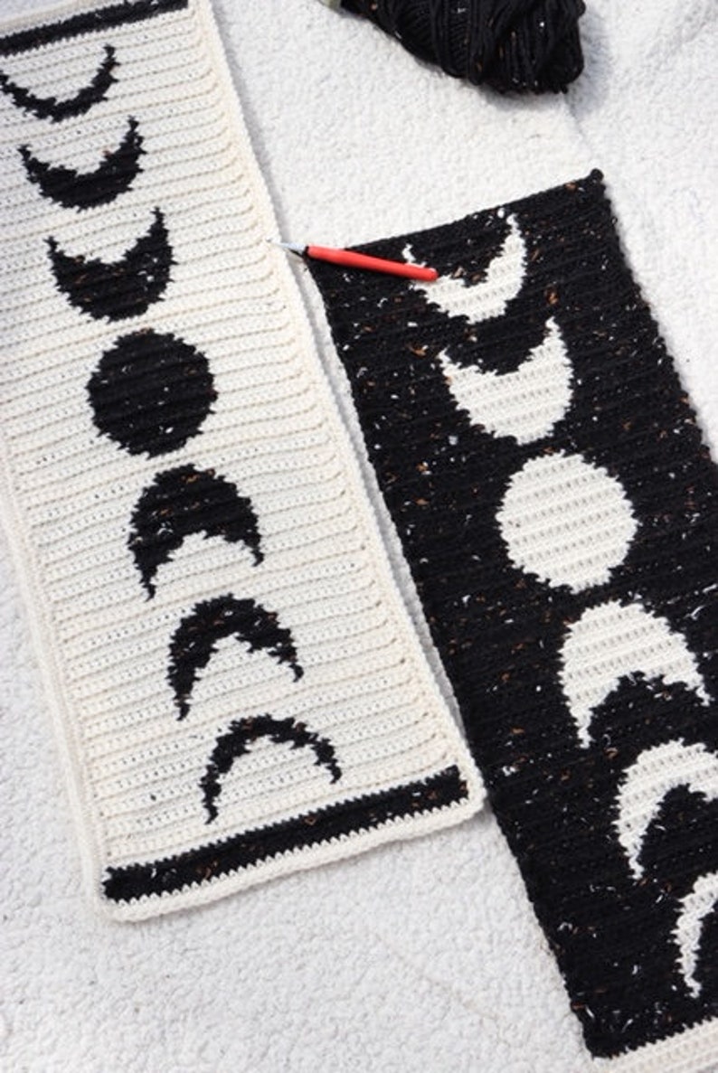 Puede incluir: Dos paneles de crochet con un dise&ntilde;o de fases lunares en blanco y negro. Los paneles est&aacute;n unidos con un gancho de crochet rojo.