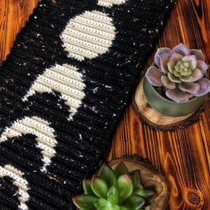 Puede incluir: Bufanda de crochet negra y blanca con un patr&oacute;n repetitivo de lunas. La bufanda est&aacute; sobre una superficie de madera con dos peque&ntilde;as suculentas verdes en macetas.