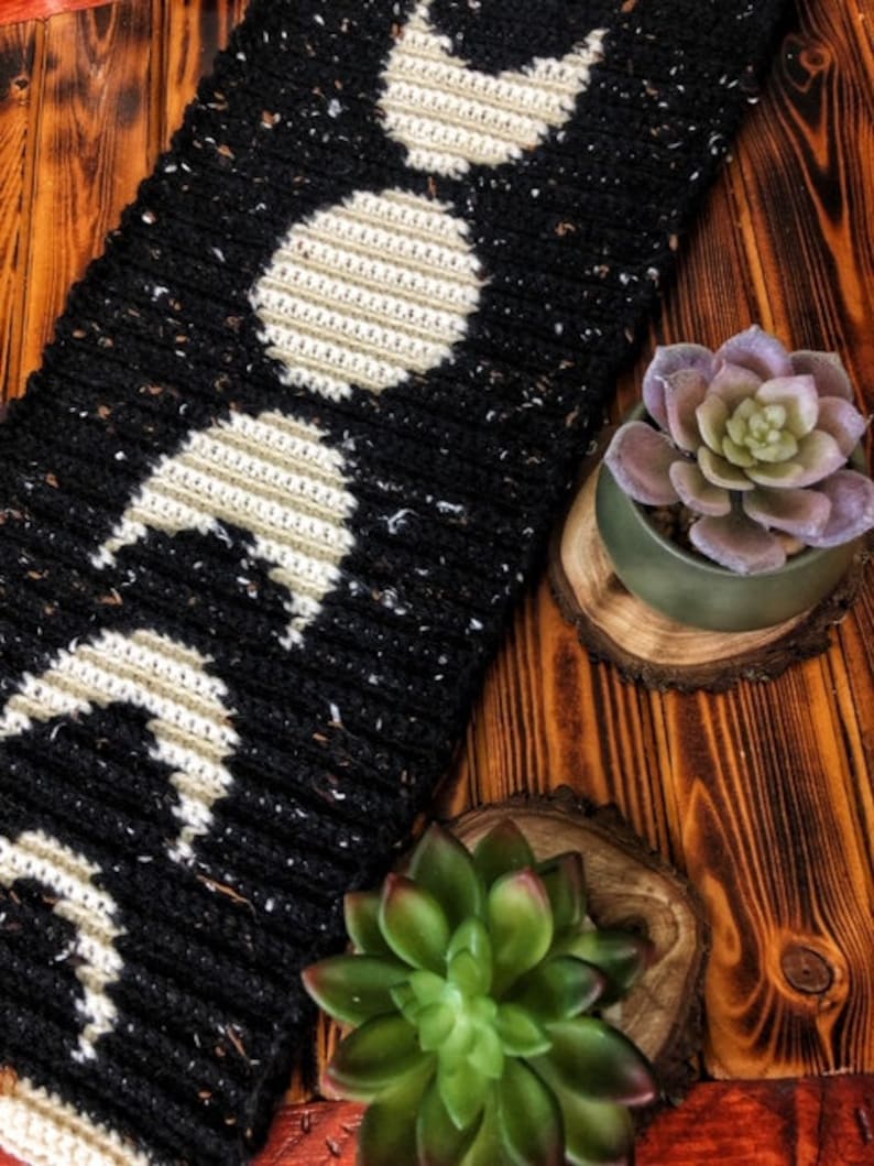 Puede incluir: Bufanda de crochet negra y blanca con un patr&oacute;n repetitivo de lunas. La bufanda est&aacute; sobre una superficie de madera con dos peque&ntilde;as suculentas verdes en macetas.