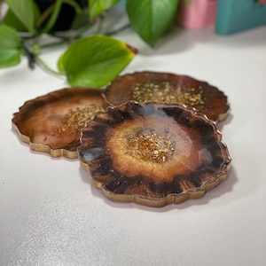 Peut inclure: Trois sous-verres en résine marron et or avec un effet marbré et des paillettes dorées. Les sous-verres ont un bord festonné.