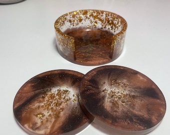 Puede incluir: Conjunto de tres posavasos de resina marrón y dorado con purpurina dorada. Los posavasos son redondos y tienen un diseño de remolino blanco. El conjunto incluye un portavasos a juego.