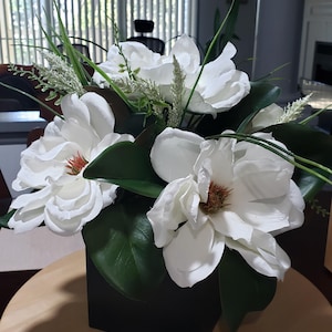 May include: A floral arrangement featuring large, white magnolia blossoms with brown centers and dark green leaves. The flowers are arranged in a square, black container, and sit on a wooden surface.