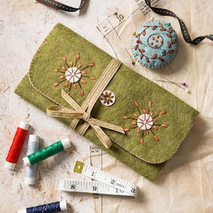 May include: An olive green felt needle case featuring embroidered starburst patterns and a beige ribbon tie. The set includes a light blue circular pincushion with floral embroidery, four spools of thread, and a white measuring tape.