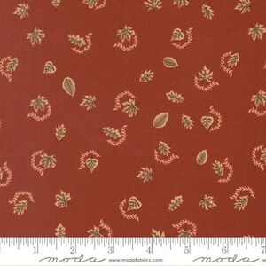 Può includere: Un tessuto color ruggine con un motivo ripetuto di piccole foglie color crema e rametti curvi. Sul bordo inferiore è visibile un righello bianco con misure in centimetri, insieme al testo moda e modafabrics.com.
