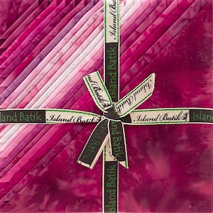 May include: A square stack of fabric squares featuring a gradient of pink and magenta batik patterns. The stack is tied with a decorative ribbon printed with the text Island Batik in a repeating pattern, secured in a bow at the centre.