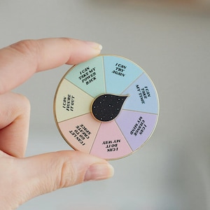 May include: A colorful, circular pin with motivational phrases like "I can take my power back" and "I can try again." The pin has a gold border and a black, star-shaped center. The pin is held by a hand.