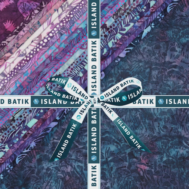 May include: A stack of folded fabric pieces featuring various purple, blue, and teal batik patterns. The fabric bundle is tied with a white ribbon printed with the text ISLAND BATIK in dark blue, arranged in a cross pattern over the textiles.