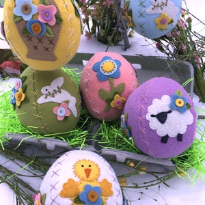 Puede incluir: Seis huevos de Pascua de fieltro decorados con diferentes diseños. Los huevos están en una caja con hierba verde. Los diseños incluyen una cesta de flores, un conejito, una flor, un cordero y un pollito.
