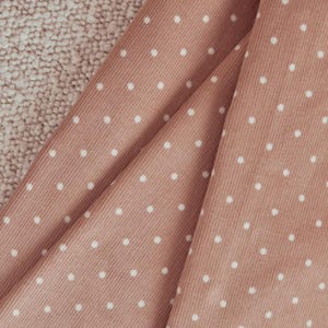 May include: A dusty rose corduroy fabric featuring a repeating pattern of small, white, evenly spaced polka dots. The material has a distinct vertical ribbed texture characteristic of corduroy, with folds creating depth and shadows across the surface.
