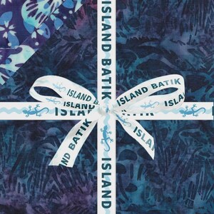 Puede incluir: Una pila de tela doblada con patrones batik en azul oscuro y morado, atada con una cinta blanca en forma de cruz. La cinta está impresa con el texto ISLAND BATIK y pequeños iconos de lagartos azules repetidos a lo largo de la cinta.