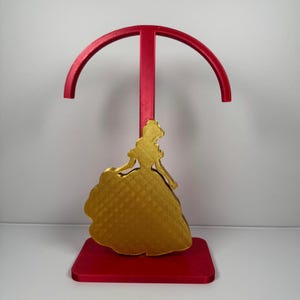 May include: A red and gold decorative stand featuring a silhouette of a woman in a ball gown. The stand has a red base and a red arch at the top. The woman's silhouette is gold.