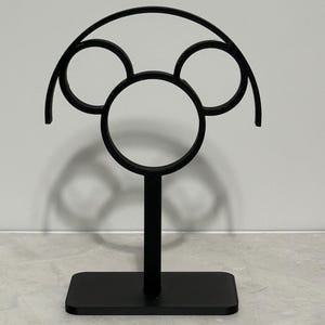 May include: A black metal decorative stand featuring a Mickey Mouse-inspired design. The stand has a rectangular base, a vertical stem, and a top section with three circular cutouts and an arched top. The stand is designed to hold items.