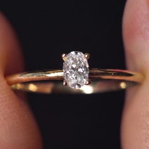 May include: A gold ring featuring a single oval-cut clear gemstone set in a four-prong setting. The band is smooth and polished, showcasing a classic design with a central stone that reflects light, highlighting the metallic luster of the gold band.