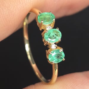 Puede incluir: Un anillo de oro con tres esmeraldas verdes de corte ovalado dispuestas en fila vertical, separadas por dos pequeñas piedras de acento claras de corte redondo engastadas en oro sobre una banda fina y pulida.