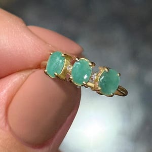 May include: A gold ring featuring three oval-cut emeralds. The emeralds are set in a row, with small accent stones between each emerald. The ring has a delicate, elegant design, perfect for a special occasion.