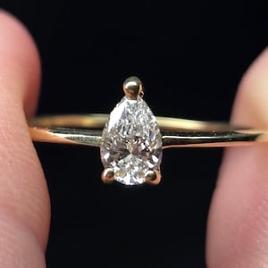 May include: A gold ring featuring a single pear-shaped, faceted clear gemstone set in a three-prong mounting. The band is thin and polished, showcasing a metallic yellow finish that contrasts with the brilliant, multi-faceted white stone.