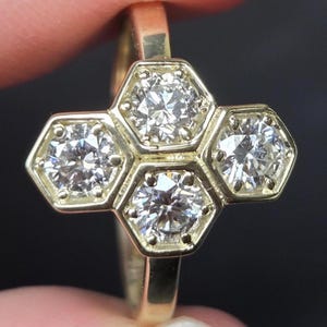 Könnte beinhalten: Ein Goldring mit einer Anordnung aus vier runden Diamanten im Brillantschliff, die jeweils in einer einzelnen sechseckigen Fassung sitzen. Die Diamanten sind geometrisch angeordnet, mit einem Stein oben, einem unten und zwei an den Seiten.