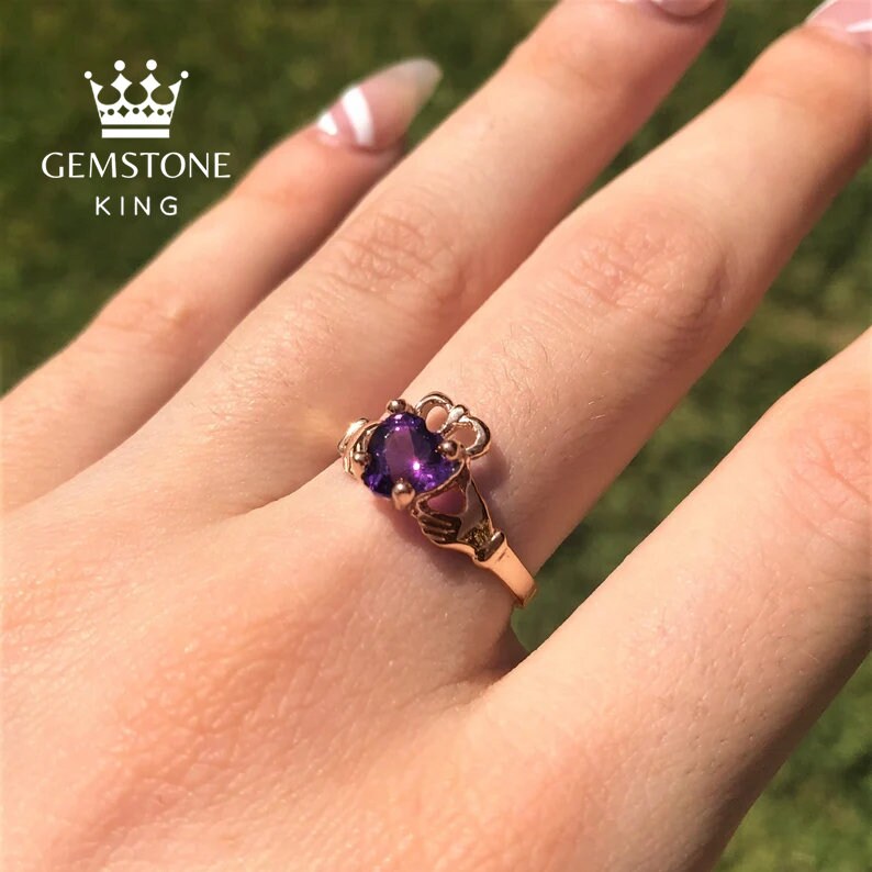 May include: A rose gold Claddagh ring with a purple heart-shaped gemstone. The ring features a crown, two hands clasped together, and a heart.