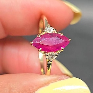 May include: A gold ring featuring a central marquise-cut vibrant pink gemstone held in a prong setting, flanked by two small round clear stones positioned above and below the main stone on a thin, polished gold band.