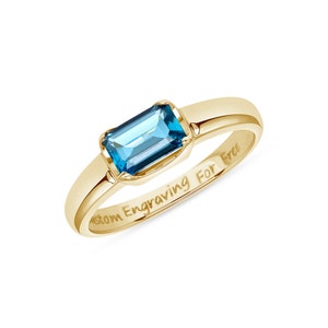 May include: A gold ring featuring a rectangular emerald-cut blue gemstone set in a polished band. The inner surface of the gold band is clearly engraved with the text Custom Engraving For Free, showcasing the smooth metal finish and setting.