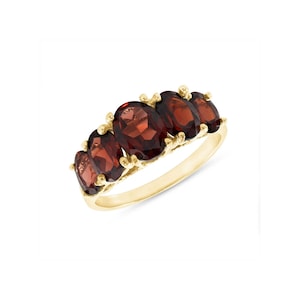 May include: A yellow gold ring featuring five oval-cut deep red garnet gemstones arranged in a row. The stones are set in a prong setting along the top of the polished gold band, creating a continuous horizontal display of dark red faceted gems.