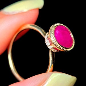 Pode incluir: Um anel de ouro com uma pedra preciosa oval de cor rosa vibrante, engastada numa moldura texturizada com contas. A lateral do engaste é acentuada com uma fila de pequenas pedras claras e brilhantes num aro de ouro liso e polido.