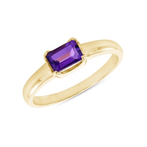 May include: A gold ring featuring a rectangular, deep purple gemstone. The ring has a simple, elegant design with a smooth, polished band. The gemstone is set in a bezel setting, enhancing its color and shape.