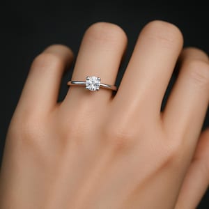 May include: A silver ring featuring a round, clear gemstone set on a slender band. The ring is on a finger, against a dark backdrop.