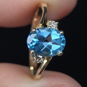 May include: A gold ring featuring a central oval-cut blue gemstone held in a four-prong setting. The band includes two small, round, clear accent stones positioned on either side of the main blue stone, creating a bypass-style design.
