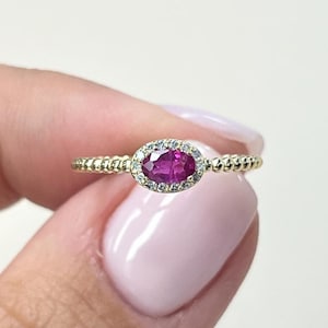 Könnte beinhalten: Ein Goldring mit einem oval geschliffenen, leuchtend pinkfarbenen Edelstein in der Mitte, der von einem Kranz aus kleinen weißen Steinen umgeben ist. Die Ringschiene hat ein strukturiertes Perlendesign, das vom Mittelstück ausgeht.