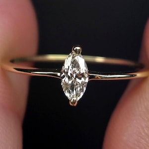 May include: A gold ring featuring a single marquise-cut clear gemstone set horizontally. The band is thin and polished, with the stone secured by two small prongs at the pointed ends. The metal has a warm yellow tone and a smooth, reflective finish.