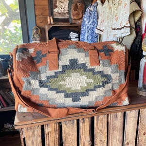 May include: A large, rectangular duffel bag with a geometric pattern in shades of orange, grey, green, and cream. The bag has brown handles and a shoulder strap, and is made of a woven fabric.