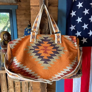 May include: Orange and white woven duffel bag with a geometric pattern. The bag has a white canvas strap and a brown leather handle.