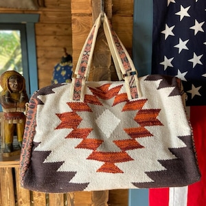 May include: A white, brown, and orange geometric patterned duffel bag with a canvas strap. The bag is hanging on a hook.