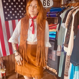 Può includere: Una donna indossa un gilet di pelle scamosciata marrone con frange, una camicetta di pizzo bianco e una gonna di pelle scamosciata marrone con frange. È in piedi davanti a un muro con una bandiera americana e un cartello che dice "66".