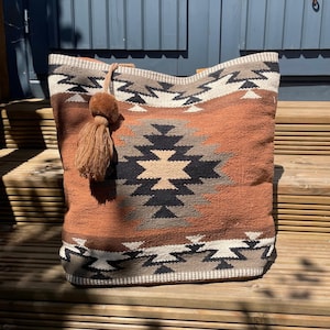 May include: A large, woven tote bag with a Southwestern geometric pattern in shades of brown, black, and cream. The bag features a decorative tassel and a sturdy handle. The bag is made of a thick, durable material, perfect for carrying items.