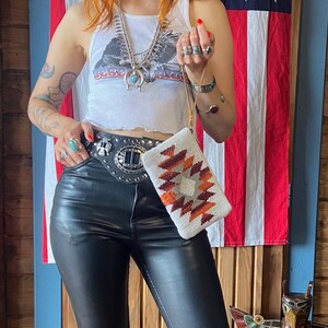 Pode incluir: Um top branco sem mangas com um gráfico de uma águia e o texto "Desert Road". A pessoa está usando um cinto de couro preto com uma fivela de prata grande e uma bolsa de mão tecida branca e laranja.