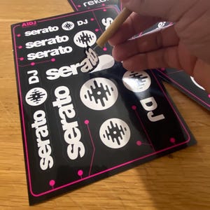 May include: Black stickers with the words "serato" and "DJ" in white, along with circular graphic designs. The stickers also feature the text "rekordbox" and the brand name "AIDJ". A wooden tool is peeling a sticker from the sheet.