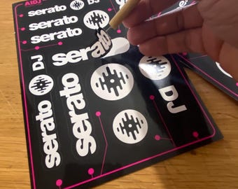 Może przedstawiać: Czarne naklejki z białymi napisami "serato" i "DJ" oraz okrągłymi wzorami graficznymi. Naklejki zawierają również tekst "rekordbox" i markę "AIDJ". Drewniane narzędzie odkleja naklejkę z arkusza.